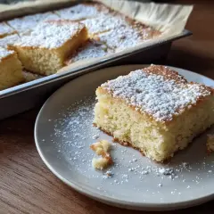En bit av den godaste kakan i långpanna, pudrad med florsocker, ligger på en tallrik. Resten av kakan syns i bakgrunden.