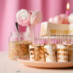 Grillade marshmallows på spett, perfekt för barnkalas fika. En tårta och strössel syns i bakgrunden.