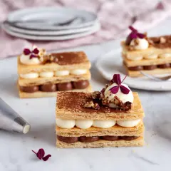Tre eleganta lager av smördeg, chokladkräm och vaniljkräm toppas med nötter och en lila blomma. En lyxig dessert redo att avnjutas.