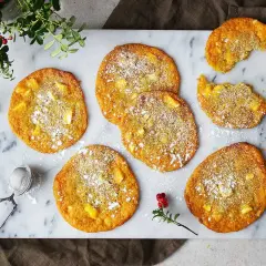 Sju gyllene saffranscookies med vit choklad, pudrade med florsocker, ligger på ett marmorfat. En bit är tagen från en cookie.