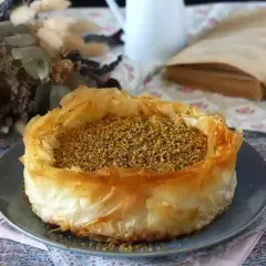 En gyllene baklava cheesecake med pistagenötter, serverad på en grå tallrik. En vacker dessert att njuta av med en god bok.