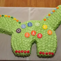 Enkel dinosaurietårta i ljusgrönt med godisdekorationer. Tårtan är formad som en dinosaurie och har bokstäver och siffror som bildar "OLIVER 2 ÅR".