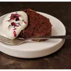 En bit saftig sockerkaka choklad med vispgrädde och blomblad på en vit tallrik med en gaffel.