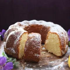 En saftig citron sockerkaka, pudrad med florsocker, serveras med citronskivor och lila blommor på en träbricka.