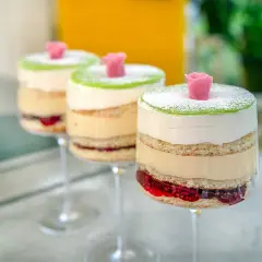 Tre vackra prinsesstårtor i glas, med lager av sockerkaka, grädde, vaniljkräm och hallonsylt, toppade med grön marsipan och en rosa ros.