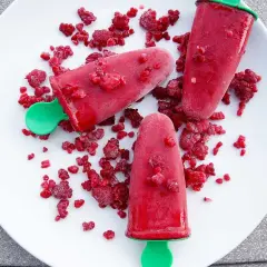Tre hallonpinnar på en vit tallrik, omgivna av frysta hallon. Ett enkelt isglass recept för sommaren.