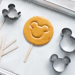 En färdiggjord dalgona cookie med Musse Pigg-form ligger på en bakplåt med flera Mickey Mouse-formar och träpinnar.