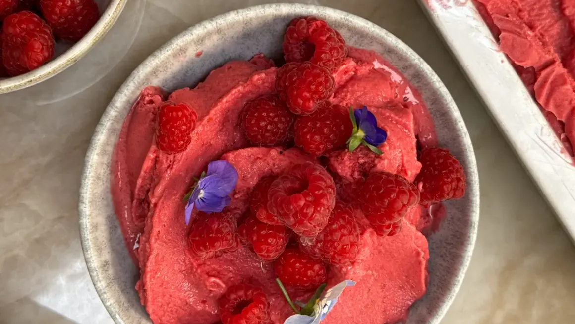 En skål med krämig hallonyoghurtglass toppad med färska hallon och små violer. En härlig sommardessert.