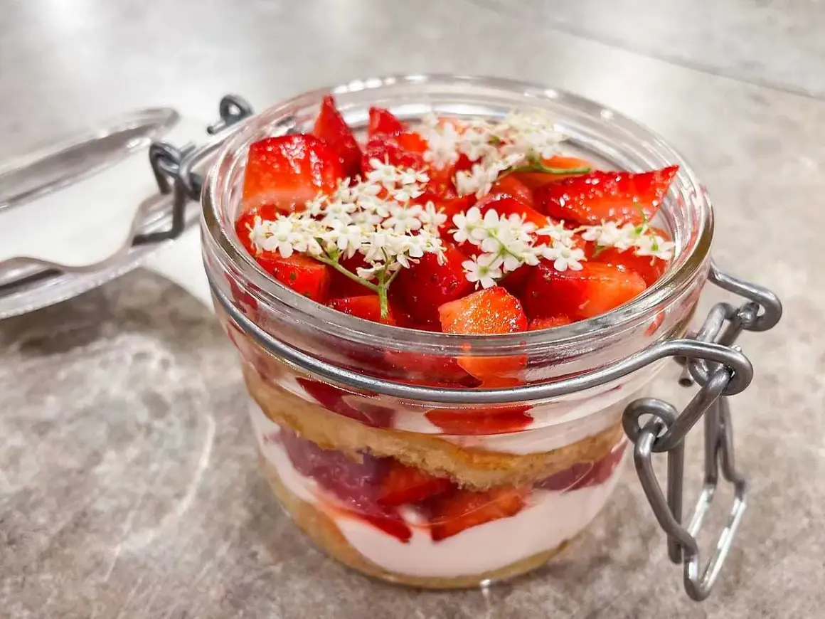 Strawberry and cream dessert layered in a glass jar, topped with fresh strawberries and small white flowers.