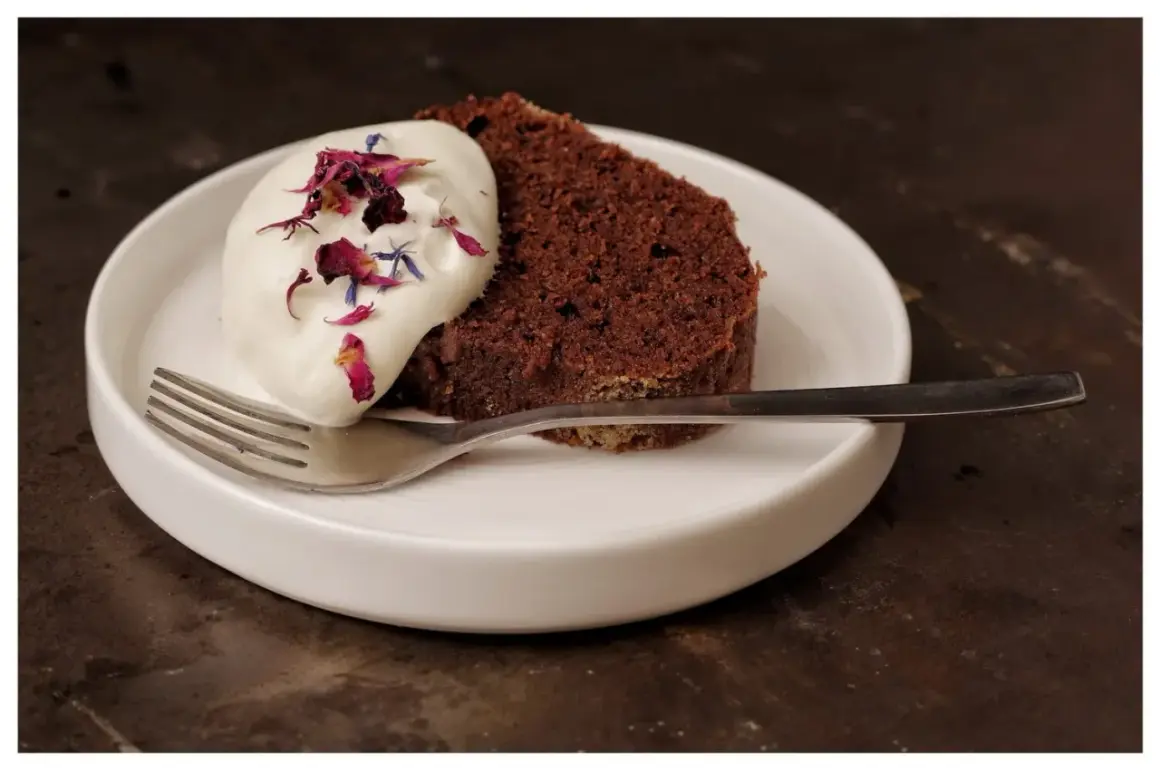 En bit saftig sockerkaka choklad med vispgrädde och blomblad på en vit tallrik med en gaffel.