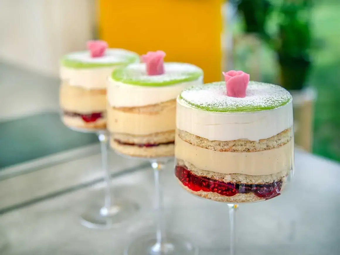 Tre vackra prinsesstårtor i glas, med lager av sockerkaka, grädde, vaniljkräm och hallonsylt, toppade med grön marsipan och en rosa ros.