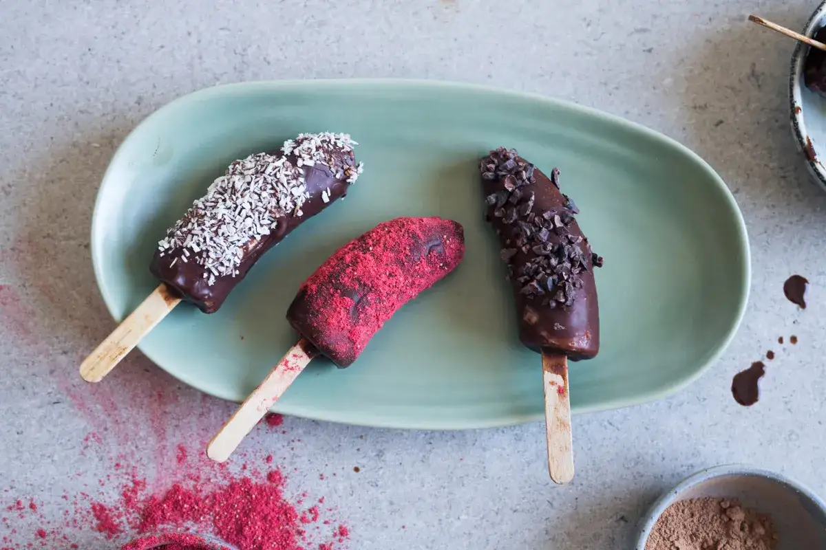 Choklad&ouml;verdragna bananer p&aring; pinne, toppade med kokos, kakaonibs och hallonpulver. Ett enkelt bananglass recept f&ouml;r en svalkande fika.