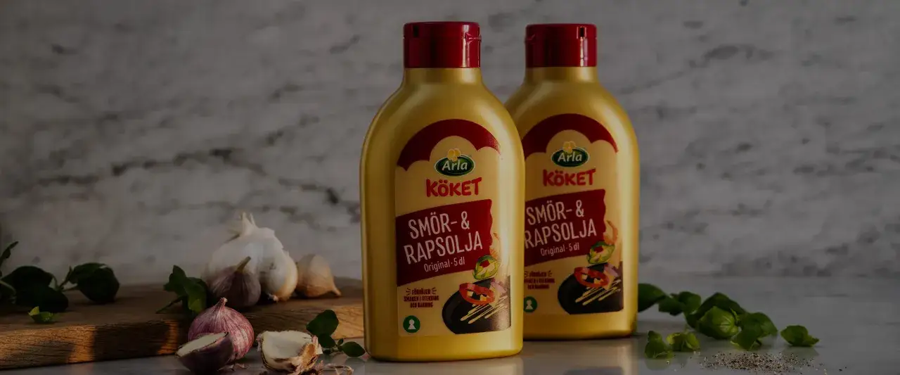 Två flaskor Arla Kökets Smör- & Rapsolja. Ett bra alternativ till smör för stekning och bakning, med vitlök och örter i bakgrunden.