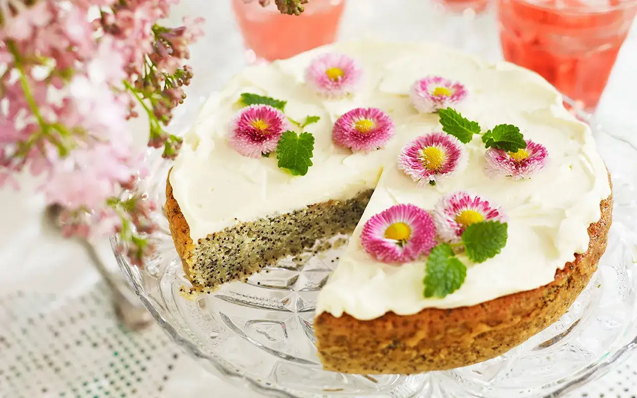 En vacker citron och vallmokaka med fluffig glasyr, dekorerad med små blommor och myntablad. Perfekt för ett somrigt fika.