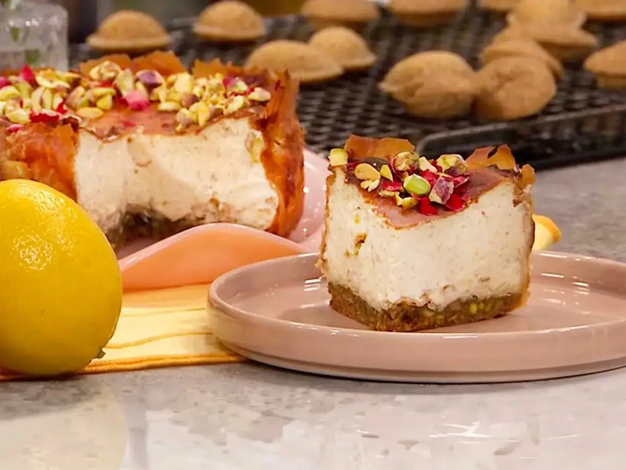 En bit baklava cheesecake med pistagenötter och rosenblad. En hel cheesecake syns i bakgrunden.