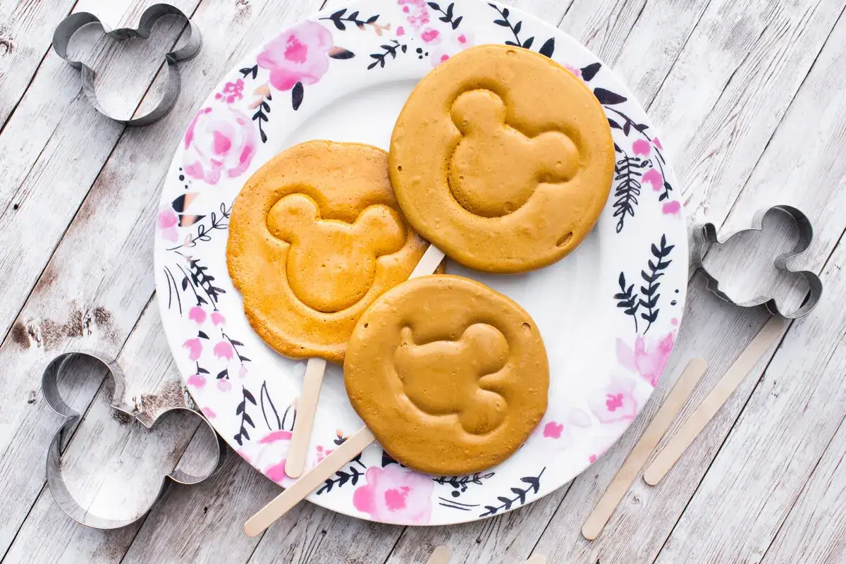 Tre s&ouml;ta dalgona cookies med Musse Pigg-motiv ligger p&aring; en blommig tallrik. Metallformar i samma motiv syns runt om.