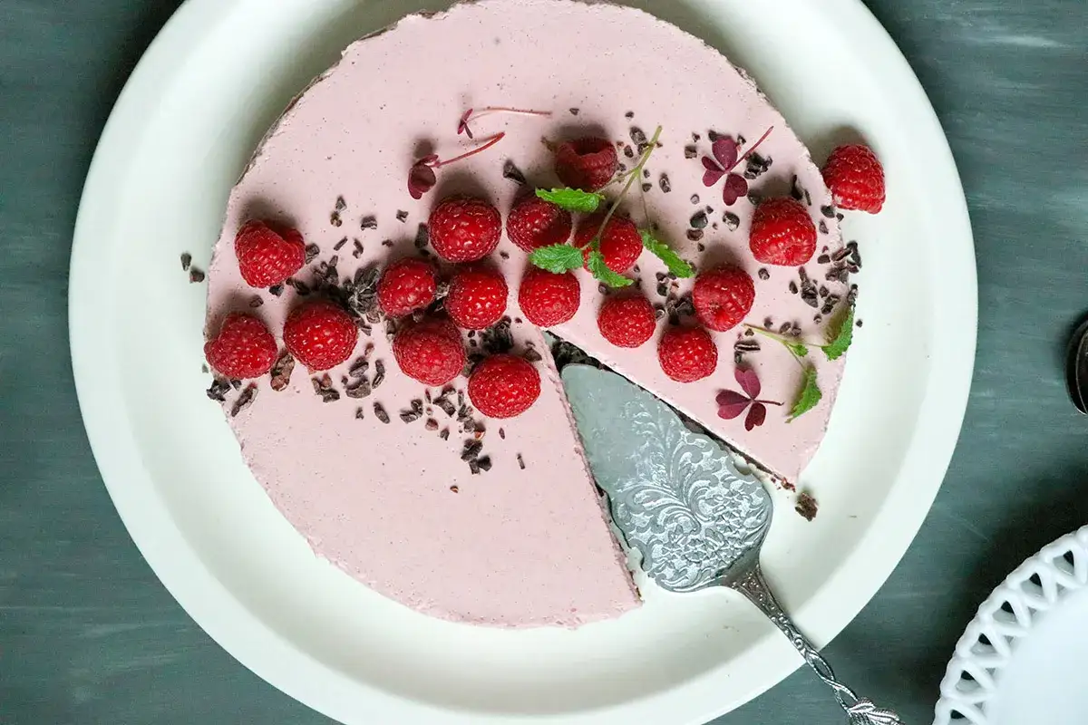 En vacker pannacotta tårta med hallon och kakaonibs. En bit är uppskuren med en elegant tårtspade.