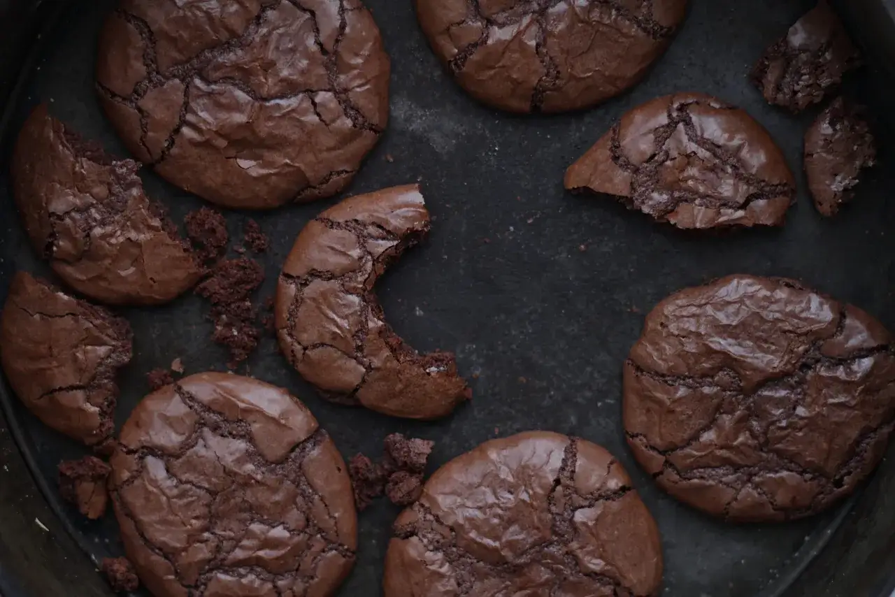 Mörka, spröda nutella kakor med sprucken yta ligger utspridda i en form. En kaka är uppäten.