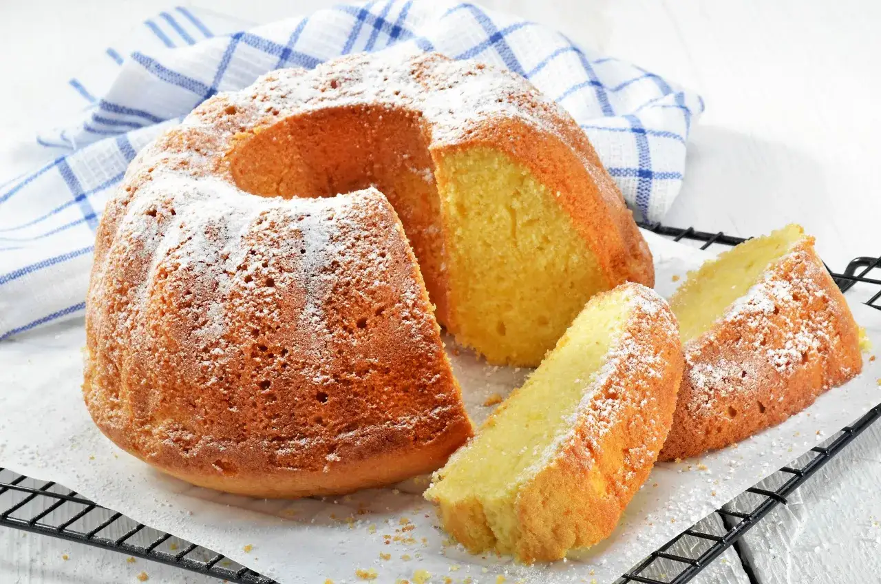 En gyllene sockerkaka med mindre socker, pudrad med florsocker, ligger på ett galler. Två skivor är upplagda bredvid.