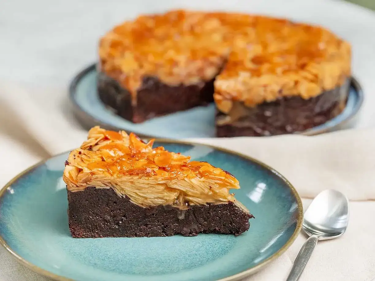En bit chokladkaka med toscatäcke av mandel. En hel toscakaka med chokladbotten syns i bakgrunden.